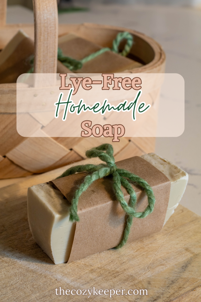 Handmade soap bar wrapped in brown paper and a piece of green yarn and sitting on a wooden cutting bar in front of a wooden basket filled with similar soap bars with text overlay that says "Lye-Free Homemade Soap."