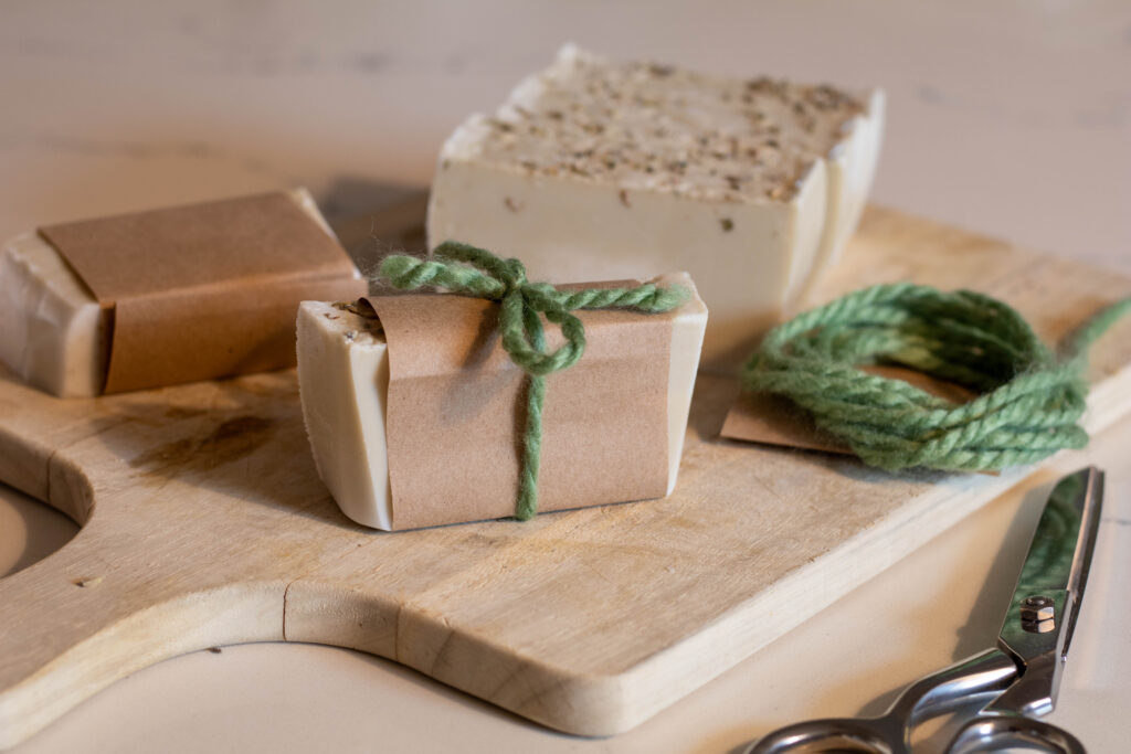 Freshly cut soap bars on a wooden cutting board 1 is wrapped in brown paper while the bar in the foreground is wrapped in brown paper and a piece of green yarn. More of the green yarn is coiled and sitting on the side next to some scissors. 