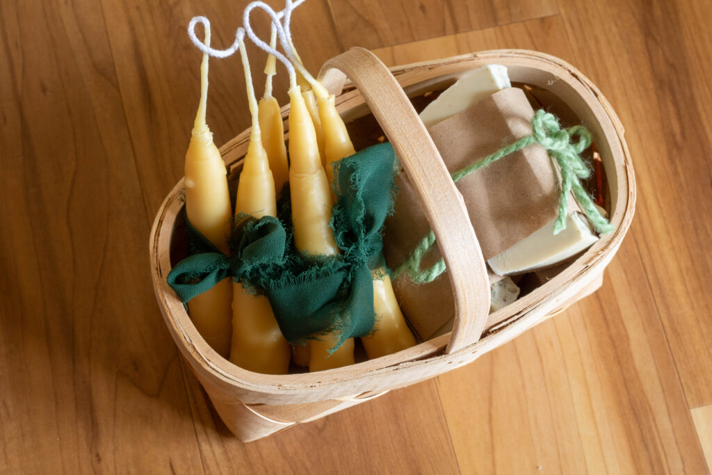Wooden basket filled with hand dipped beeswax candles and homemade lye-free soap wrapped in brown paper and green yarn. 