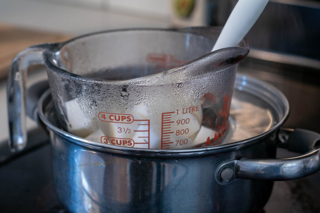 Large glass measuring cup filled with melt and pour soap base sitting in a stainless steel pot of boiling water. 