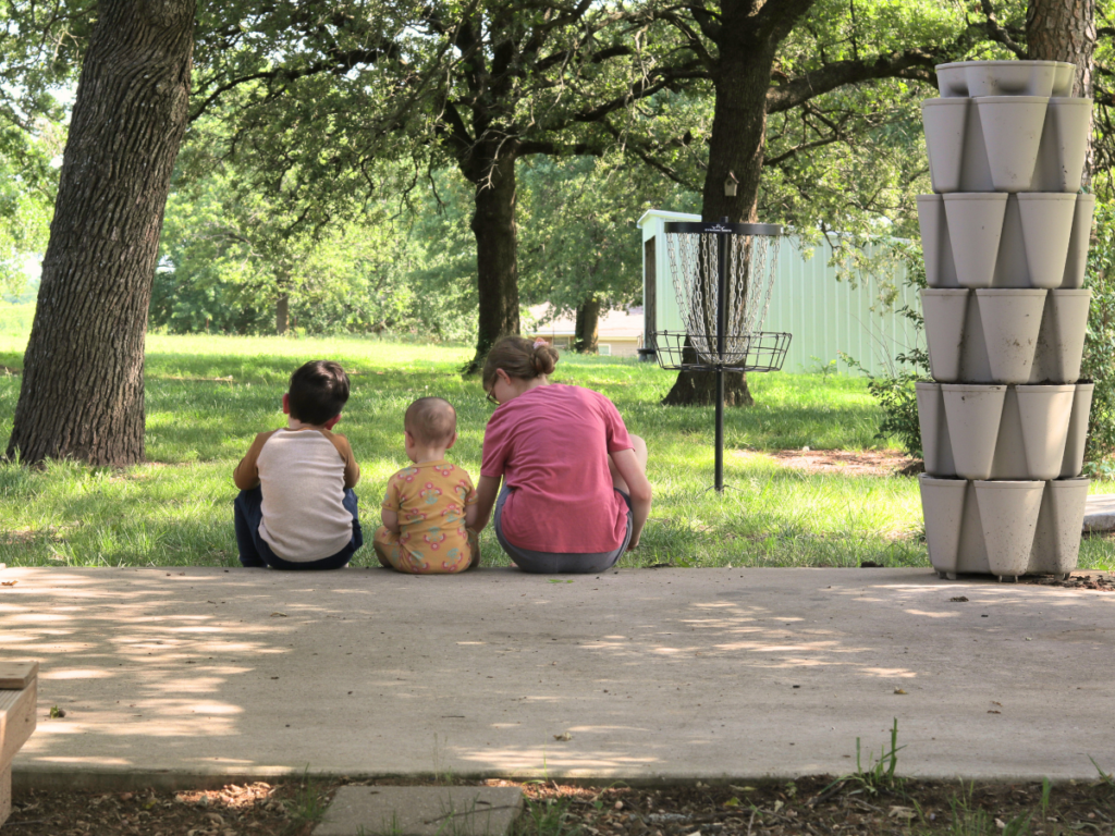 3 kids sitting on a cement slab outside with mature trees and green grass in the background and a greenstalk vertical planter to their right.