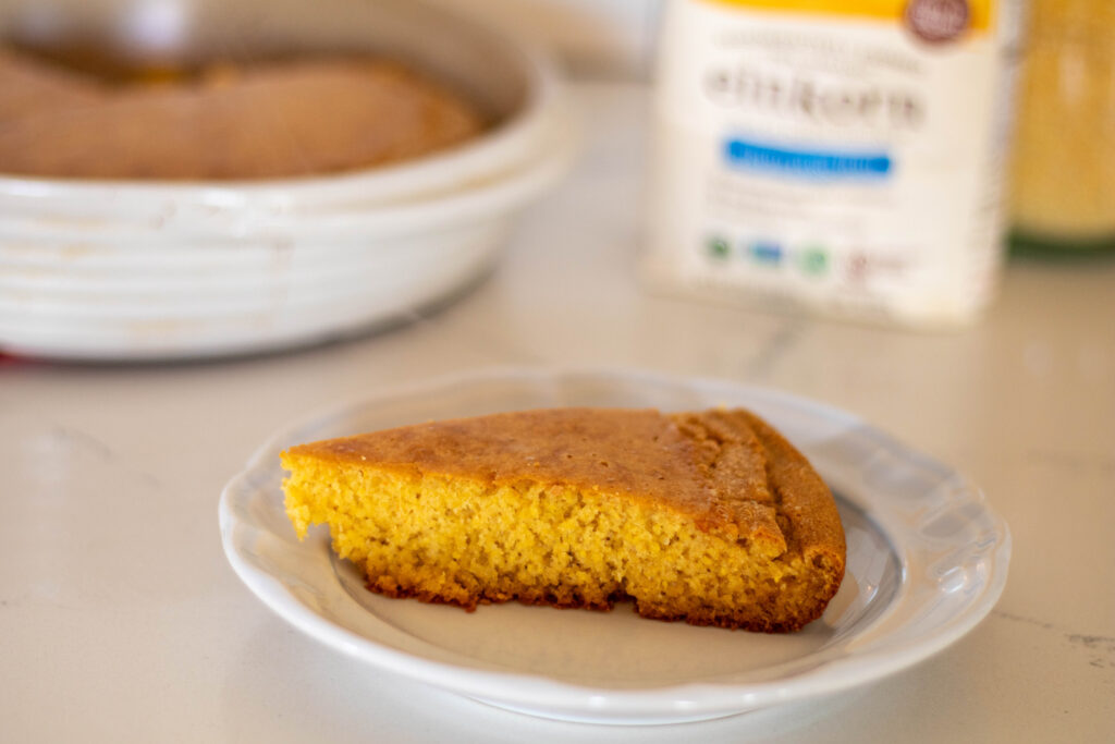 Slice of einkorn cornbread on a plate with pan of einkorn cornbread in the background with other ingredients. 
