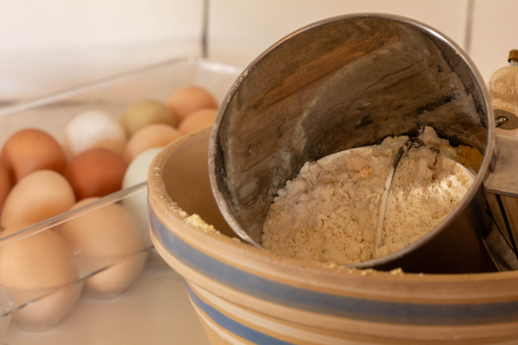 Vintage stone bowl with sifter inside filled with dry einkorn cornbread ingredients and a container of farm fresh eggs next to it.