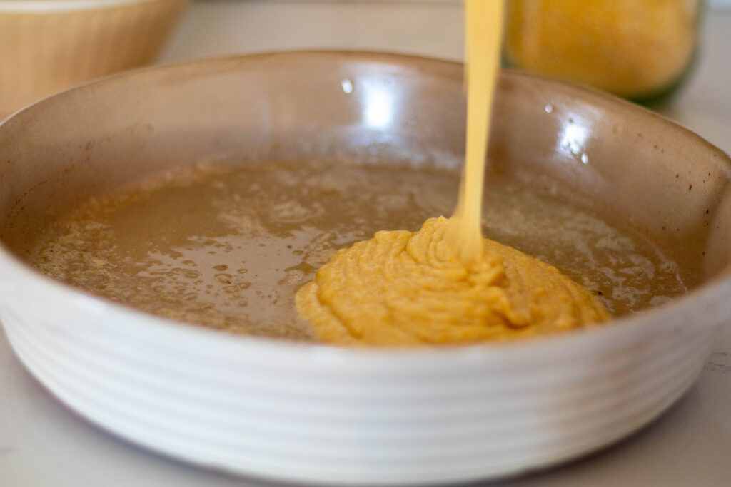 Einkorn cornbread batter being poured into buttered round stoneware pan.