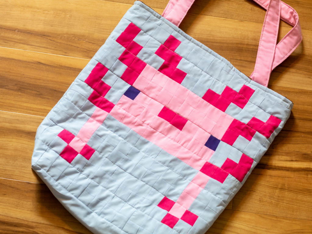 Pixel art quilted Minecraft Axolotl tote bag lying on wooden background.