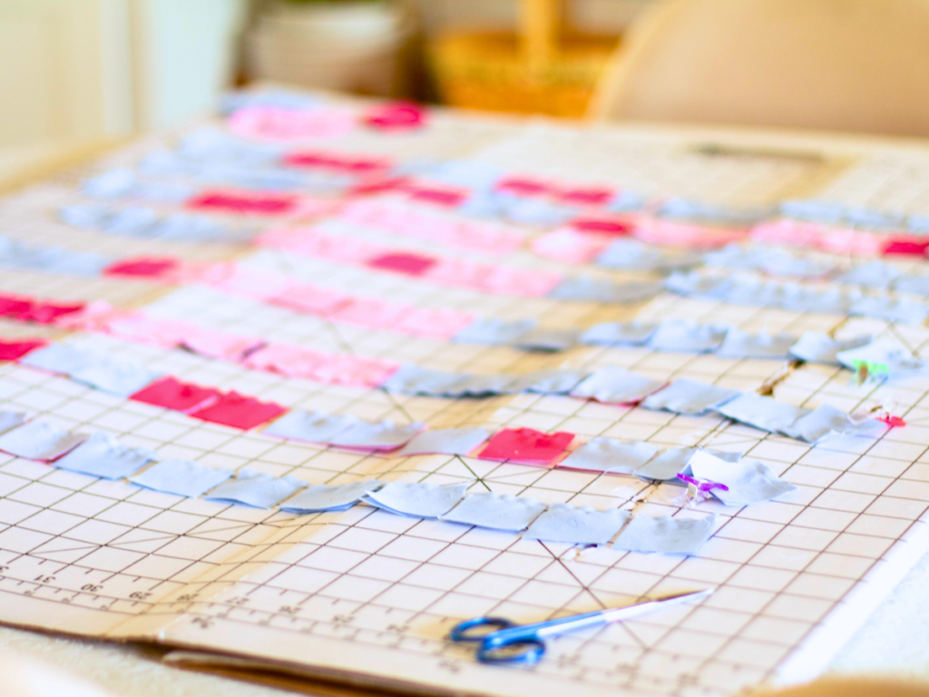 Folded over pixel art quilting squares laying on a gridded ruler background with thread scissors lying near by.
